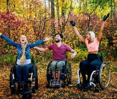 Three adults in wheelchairs with their arms in the air in a fall outdoor setting.